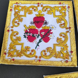 May include: A square white fabric panel featuring gold thread embroidery in a scrolling border pattern. The center displays three red hearts, one with a crown of thorns, one pierced by a gold dagger, and one adorned with white flowers and green leaves.