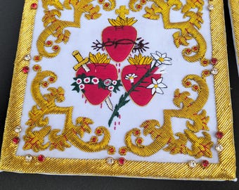 May include: A square white fabric panel featuring gold thread embroidery in a scrolling border pattern. The center displays three red hearts, one with a crown of thorns, one pierced by a gold dagger, and one adorned with white flowers and green leaves.