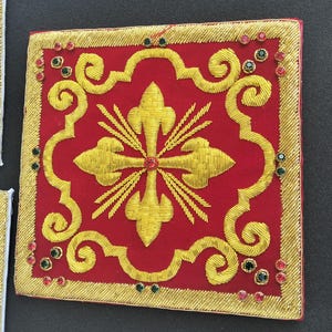 Può includere: Una toppa quadrata in tessuto rosso con un intricato ricamo in filo dorato raffigurante un motivo centrale a croce gigliata, circondato da un bordo decorativo dorato. Il design è arricchito da piccoli strass circolari rossi e verdi sparsi.