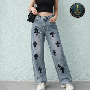 Puede incluir: Vaqueros de mezclilla de color claro con aplicaciones de cruces negras. Los vaqueros tienen un corte relajado y un diseño clásico de cinco bolsillos. Las cruces están espaciadas uniformemente en la parte delantera de los vaqueros. El modelo lleva un top negro y zapatillas blancas.