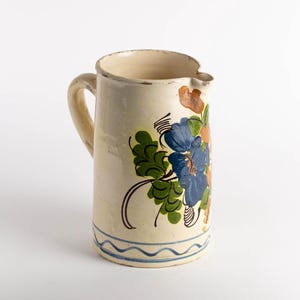 May include: Cream-colored ceramic pitcher with a handle and spout. The pitcher is decorated with blue, orange, and green floral designs. A blue wavy line borders the base.