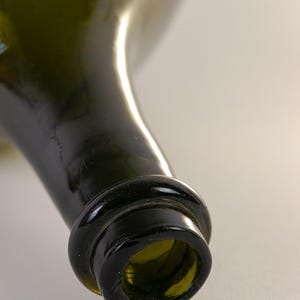 May include: Close-up of a green glass bottle. The bottle's neck and opening are in focus, showcasing a dark green hue. The rim of the bottle is thick and rounded, with a slight texture. The background is a soft, neutral color.