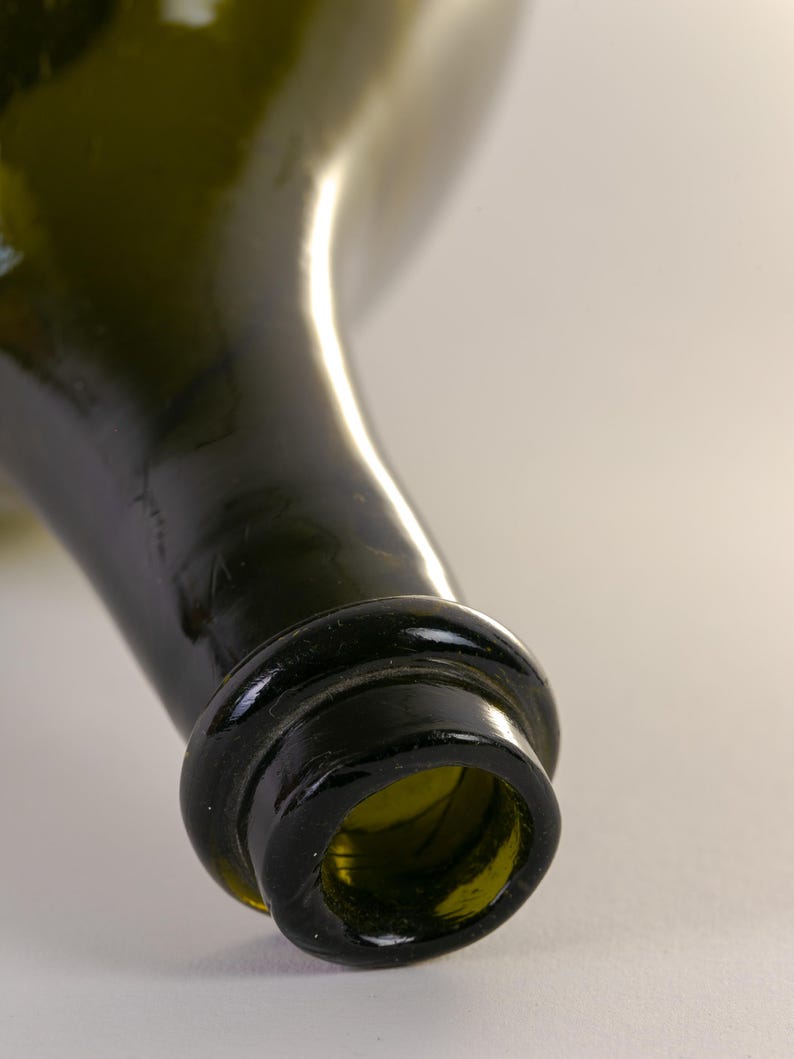 May include: Close-up of a green glass bottle. The bottle's neck and opening are in focus, showcasing a dark green hue. The rim of the bottle is thick and rounded, with a slight texture. The background is a soft, neutral color.