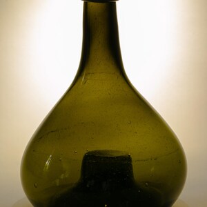 May include: A vintage olive green glass bottle with a rounded body and a long neck. The bottle has a dark green base and a small, rounded lip. The glass has a slightly textured surface with small bubbles.