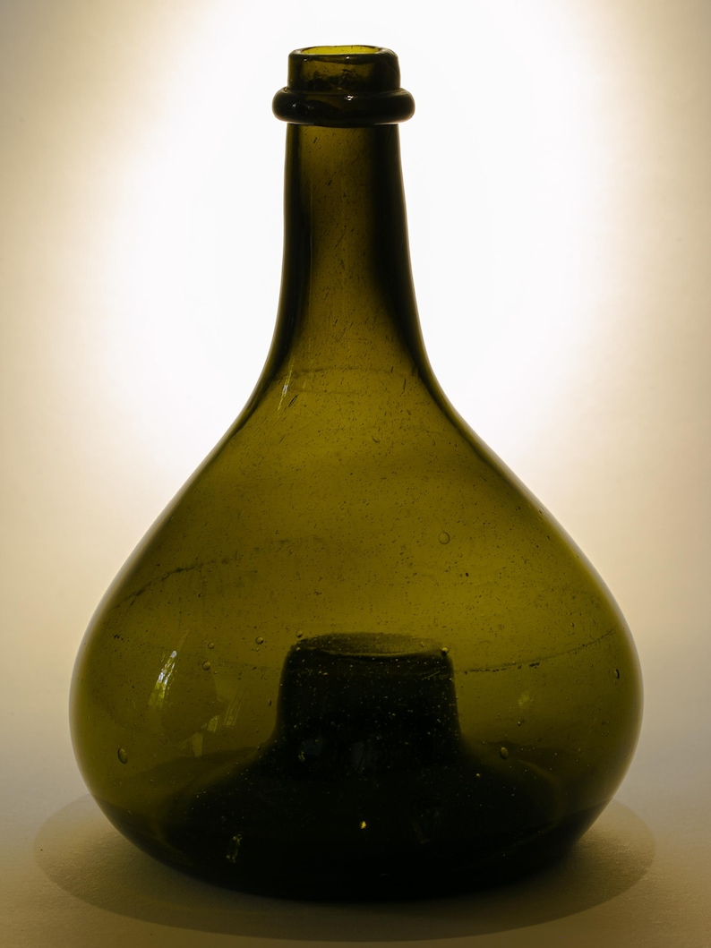May include: A vintage olive green glass bottle with a rounded body and a long neck. The bottle has a dark green base and a small, rounded lip. The glass has a slightly textured surface with small bubbles.