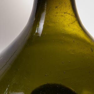 May include: Close-up of a green glass bottle with a rounded body and a long neck. The bottle has a slightly textured surface with small bubbles and imperfections, suggesting it is handmade. The glass has a subtle olive-green hue.