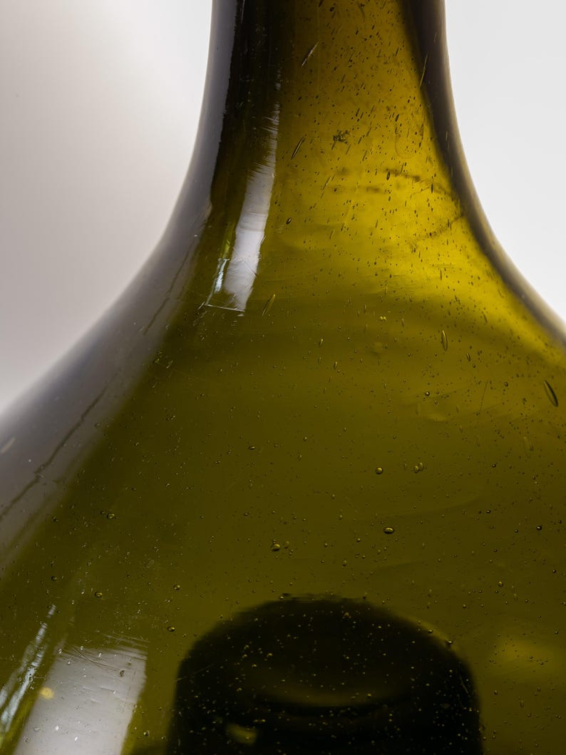 May include: Close-up of a green glass bottle with a rounded body and a long neck. The bottle has a slightly textured surface with small bubbles and imperfections, suggesting it is handmade. The glass has a subtle olive-green hue.