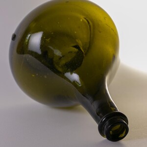 May include: An antique olive green glass bottle with a rounded body and a long, narrow neck. The bottle has a dark, textured rim and a slightly irregular shape, suggesting it is hand-blown. The glass has a slight greenish tint.