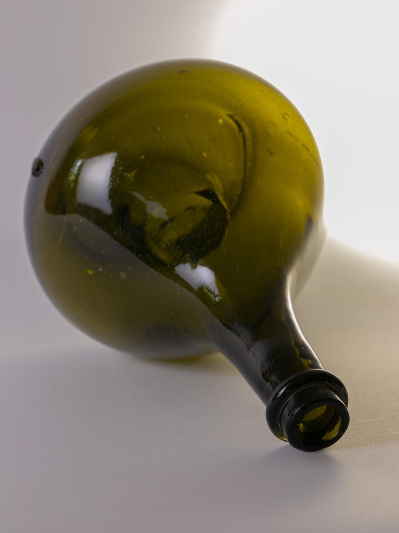 May include: An antique olive green glass bottle with a rounded body and a long, narrow neck. The bottle has a dark, textured rim and a slightly irregular shape, suggesting it is hand-blown. The glass has a slight greenish tint.