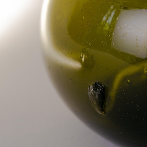 May include: Close-up of a green glass bottle with a rounded shape. The glass has a glossy finish with small bubbles and a dark, irregular inclusion. The background is a soft, neutral color.