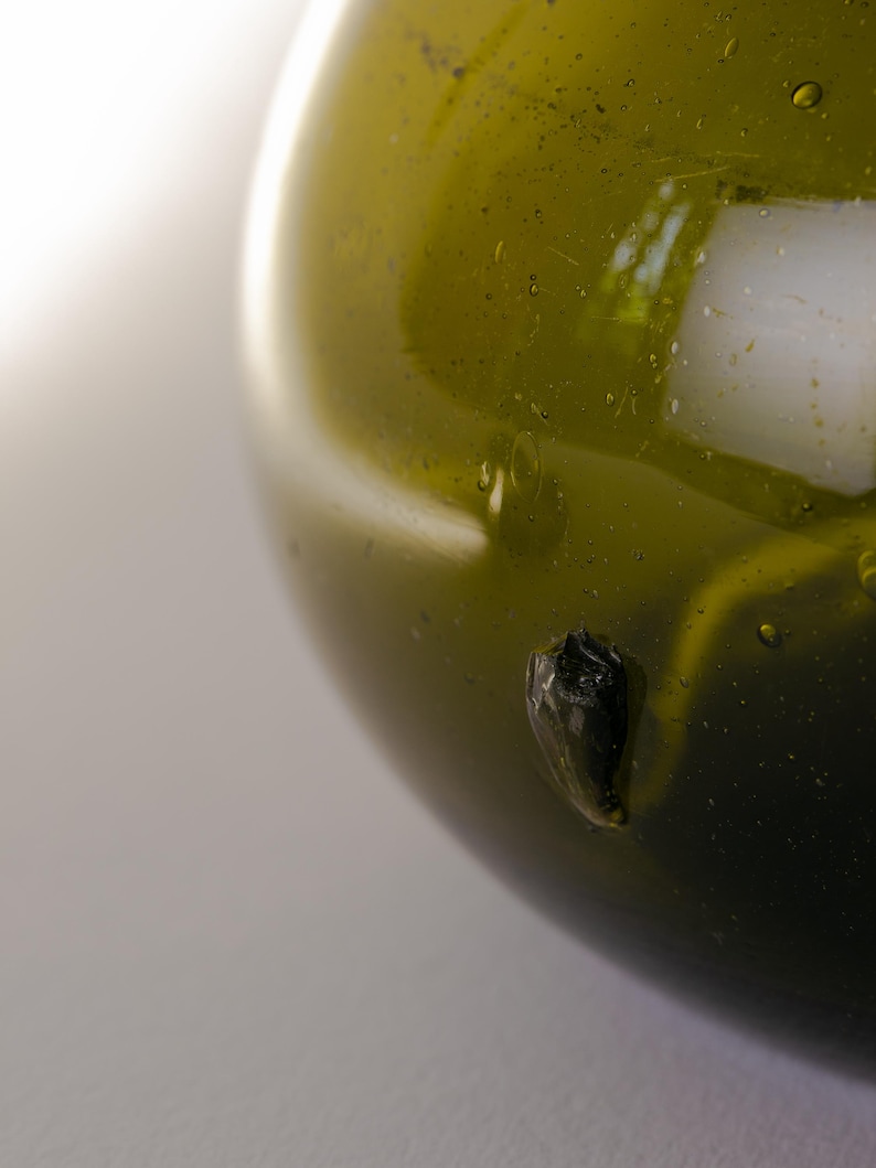 May include: Close-up of a green glass bottle with a rounded shape. The glass has a glossy finish with small bubbles and a dark, irregular inclusion. The background is a soft, neutral color.