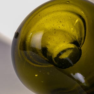 May include: Close-up of a green glass bottle with a rounded body and a long, slender neck. The glass has a slightly textured surface with small bubbles. The bottle is olive green in color and appears to be vintage.