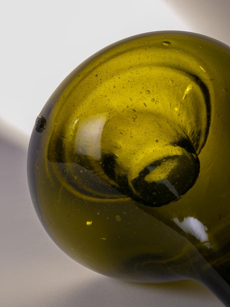 May include: Close-up of a green glass bottle with a rounded body and a long, slender neck. The glass has a slightly textured surface with small bubbles. The bottle is olive green in color and appears to be vintage.