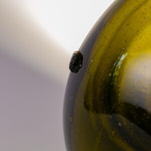 May include: Close-up of a green glass bottle with a dark, textured spot on its side. The bottle has a rounded shape and a slightly uneven surface, with visible imperfections. The glass has a subtle, yellowish-green hue.