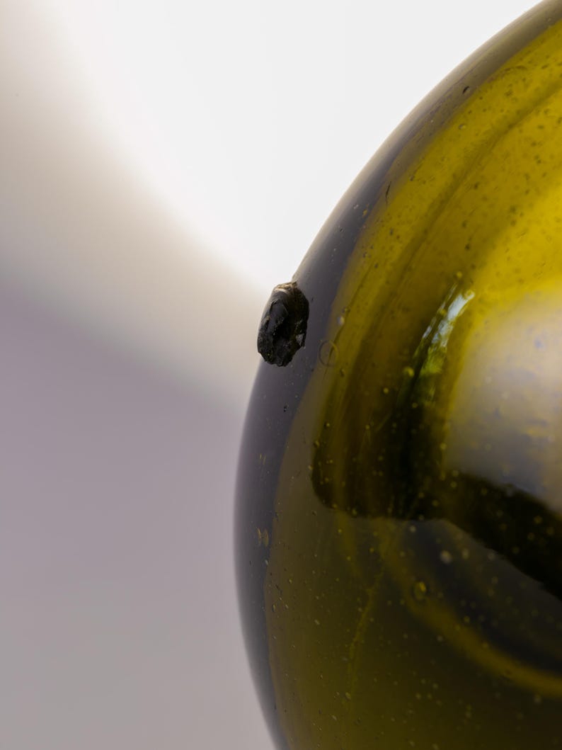 May include: Close-up of a green glass bottle with a dark, textured spot on its side. The bottle has a rounded shape and a slightly uneven surface, with visible imperfections. The glass has a subtle, yellowish-green hue.
