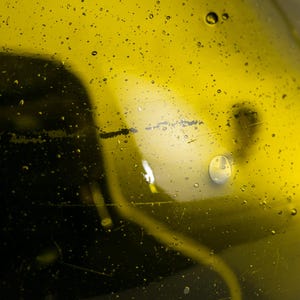May include: Close-up of a clear glass bottle filled with a yellow liquid, possibly oil. Small water droplets are scattered across the surface. The background is a darker shade of yellow, with a blurred shape.