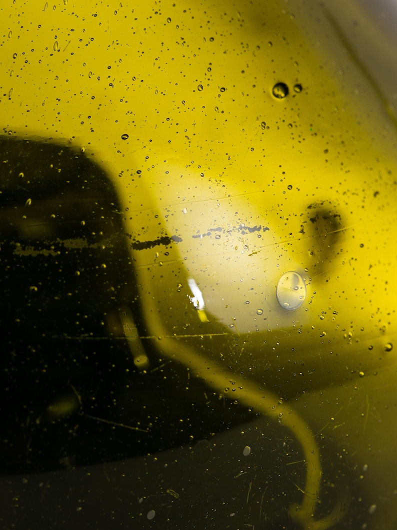 May include: Close-up of a clear glass bottle filled with a yellow liquid, possibly oil. Small water droplets are scattered across the surface. The background is a darker shade of yellow, with a blurred shape.