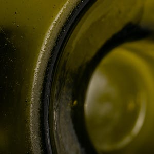 May include: Close-up of a green glass bottle's rim. The bottle's opening is visible, with a dark, rounded edge. The glass has a slightly textured surface, and the interior is a lighter shade of green.