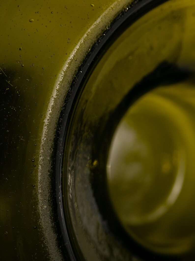 May include: Close-up of a green glass bottle's rim. The bottle's opening is visible, with a dark, rounded edge. The glass has a slightly textured surface, and the interior is a lighter shade of green.