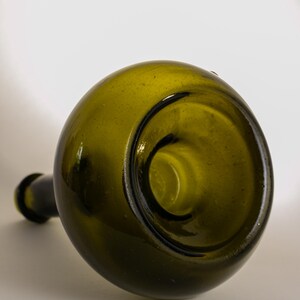 May include: An antique green glass bottle with a rounded body and a long, narrow neck. The bottle is lying on its side, showcasing its unique shape and the dark interior of the opening. The glass has a slightly textured surface.
