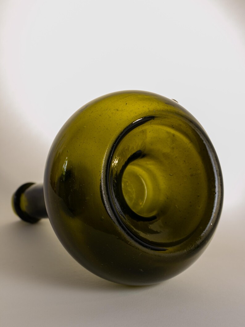 May include: An antique green glass bottle with a rounded body and a long, narrow neck. The bottle is lying on its side, showcasing its unique shape and the dark interior of the opening. The glass has a slightly textured surface.