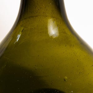 May include: Close-up of a green glass bottle with a wide base and a narrow neck. The bottle has a slightly textured surface with small bubbles and imperfections. The glass has a vintage appearance.