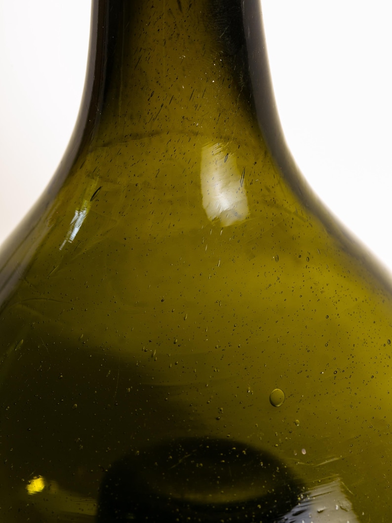 May include: Close-up of a green glass bottle with a wide base and a narrow neck. The bottle has a slightly textured surface with small bubbles and imperfections. The glass has a vintage appearance.