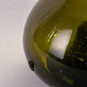 May include: Close-up of a green glass bottle with a rounded shape. The bottle has a dark green hue and a smooth, reflective surface. Small bubbles are visible within the glass. A dark, textured spot is on the side of the bottle.