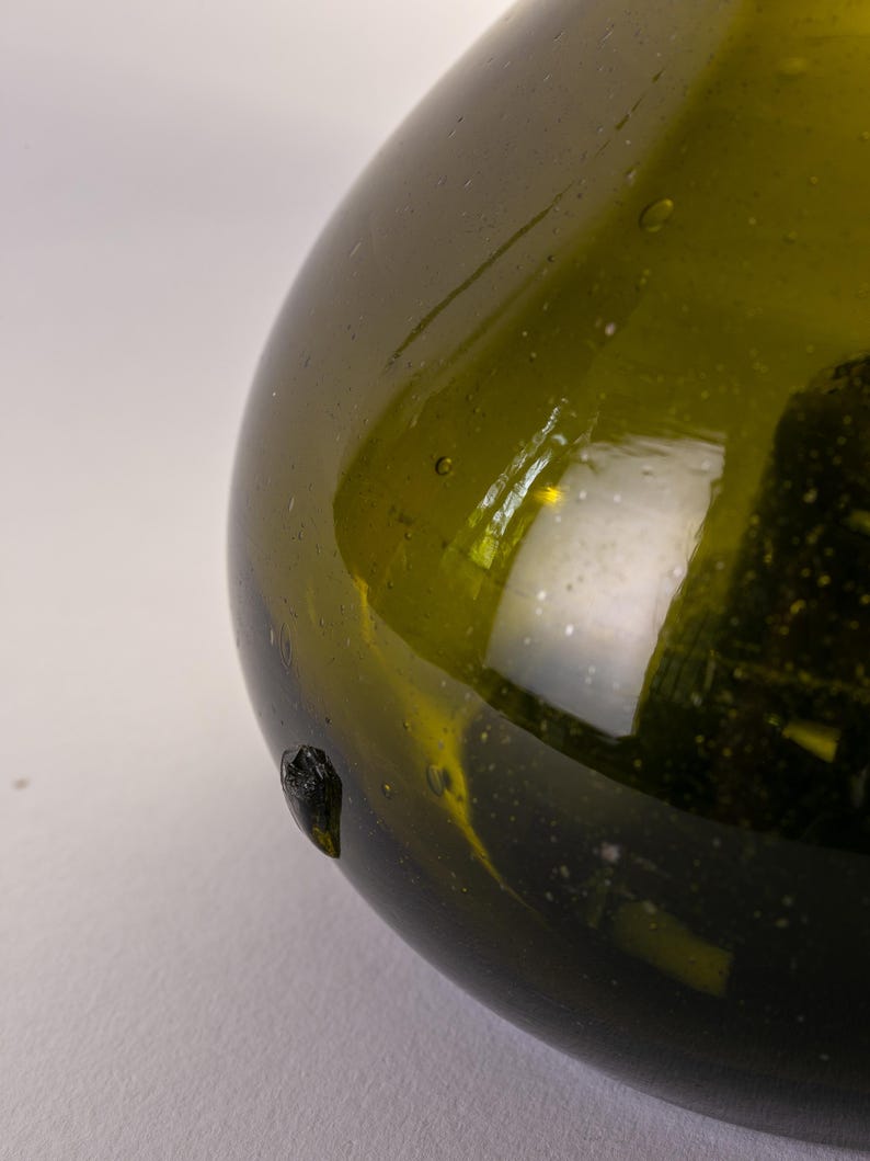 May include: Close-up of a green glass bottle with a rounded shape. The bottle has a dark green hue and a smooth, reflective surface. Small bubbles are visible within the glass. A dark, textured spot is on the side of the bottle.