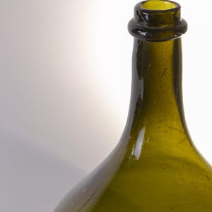 May include: A green glass bottle with a rounded body and a narrow neck. The bottle has a dark green color and a slightly textured surface. The neck of the bottle has a ring around it. The bottle is empty.