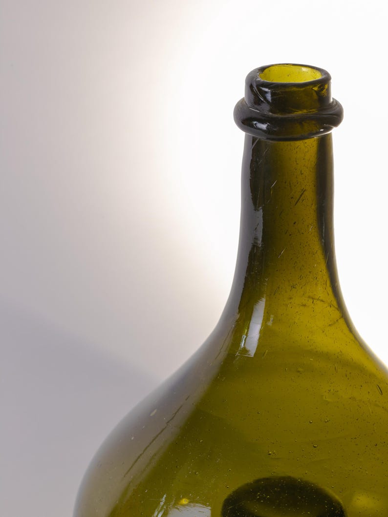 May include: A green glass bottle with a rounded body and a narrow neck. The bottle has a dark green color and a slightly textured surface. The neck of the bottle has a ring around it. The bottle is empty.