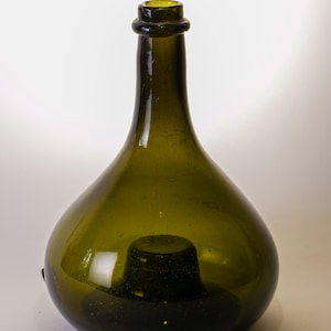 May include: A green glass bottle with a rounded body and a narrow neck. The bottle has a dark green hue and a slightly textured surface. The bottle's opening is reinforced with a dark ring.