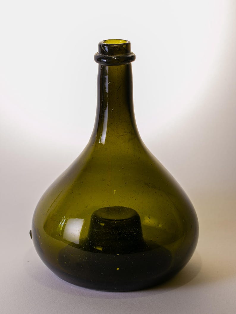 May include: A green glass bottle with a rounded body and a narrow neck. The bottle has a dark green hue and a slightly textured surface. The bottle's opening is reinforced with a dark ring.