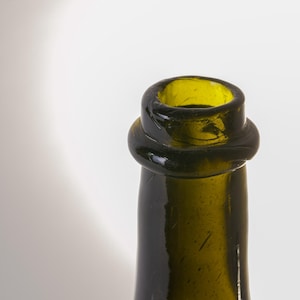 May include: Close-up of a green glass bottle neck. The bottle has a rounded lip and a slightly bulbous ring around the neck. The glass has a slightly textured surface and a dark green color.