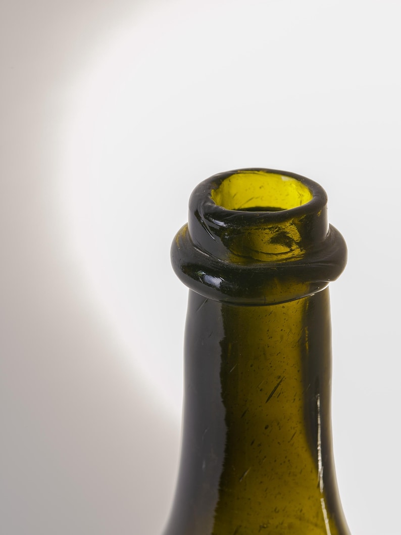 May include: Close-up of a green glass bottle neck. The bottle has a rounded lip and a slightly bulbous ring around the neck. The glass has a slightly textured surface and a dark green color.