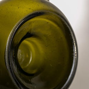 May include: Close-up of the opening of a green glass bottle. The bottle's interior is visible, showing a smooth, rounded shape. The glass has a slightly textured surface and a dark rim.