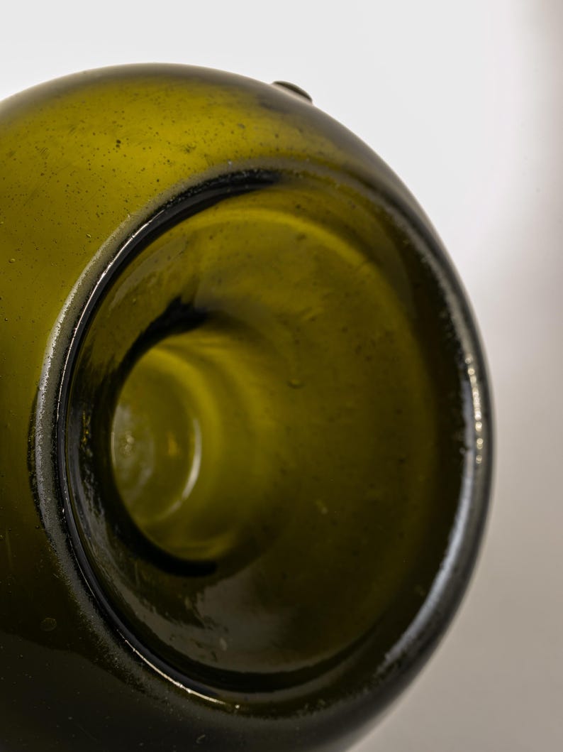 May include: Close-up of the opening of a green glass bottle. The bottle's interior is visible, showing a smooth, rounded shape. The glass has a slightly textured surface and a dark rim.