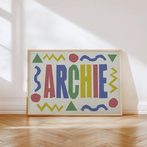 May include: A framed print featuring the name "ARCHIE" in large, colorful letters. The letters are in red, blue, yellow, and green. The print is decorated with geometric shapes in a playful design, set against a neutral background.