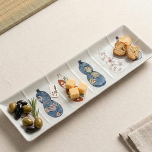 Puede incluir: Plato rectangular de cerámica blanca con un diseño de calabaza pintado a mano en azul, rosa y blanco. El plato está dividido en secciones y contiene aceitunas, cubos de queso y galletas saladas. También se ve una pequeña etiqueta con texto japonés.
