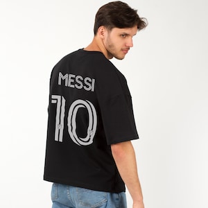 以下が含まれることがあります： 背中に白で「MESSI」の名前と「10」の数字がプリントされた黒の半袖Tシャツ。 ゆったりとしたフィット感で、柔らかい素材で作られています。