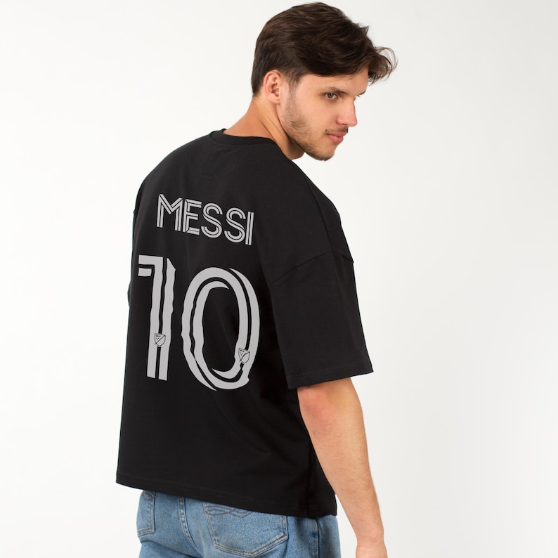 以下が含まれることがあります： 背中に白で「MESSI」の名前と「10」の数字がプリントされた黒の半袖Tシャツ。 ゆったりとしたフィット感で、柔らかい素材で作られています。