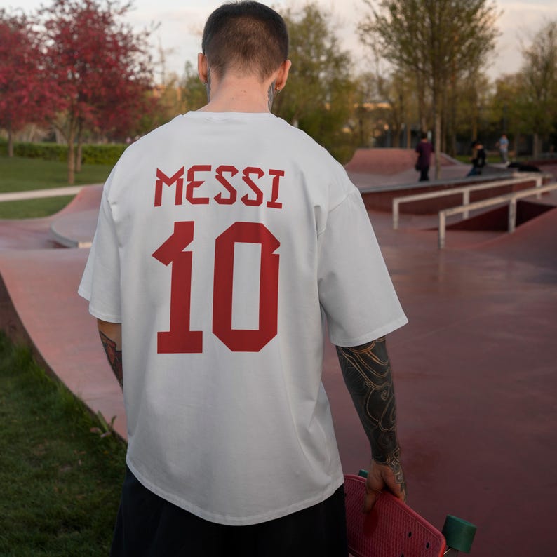 以下が含まれることがあります： 背中に赤で「MESSI」という名前と数字の「10」がプリントされた白いTシャツ。 ピンクのスケートボードを持った人が、スケートパークでそのシャツを着ています。