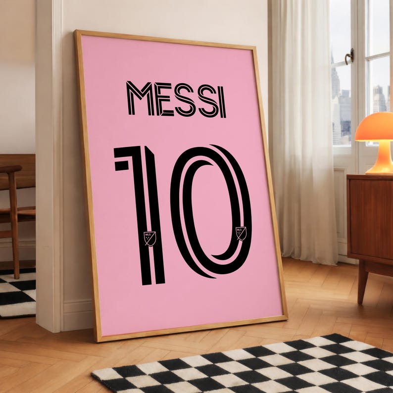 以下が含まれることがあります： 黒い文字で「MESSI」と書かれたピンク色のポスター。その下に黒い数字の「10」が描かれています。数字は白い縁取りと2つの小さなロゴが付いています。フレームは薄茶色です。ポスターは白い壁に立てかけられています。