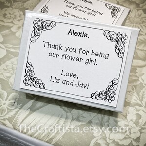 May include: White gift box with a black and white decorative border. The box has a thank you message for a flower girl named Alexis. The message reads "Alexis, Thank you for being our flower girl. Love, Liz and Javi".