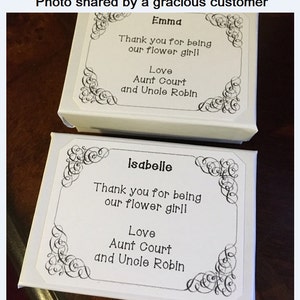 May include: Two white gift boxes with black decorative borders and text. The top box says "Emma Thank you for being our flower girl! Love Aunt Court and Uncle Robin". The bottom box says "Isabelle Thank you for being our flower girl! Love Aunt Court and Uncle Robin".