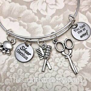 May include: Silver charm bracelet with five charms: a paw print, a comb and brush, a pair of scissors, a charm that says "Shear Genius", and a charm that says "You had me at woof".