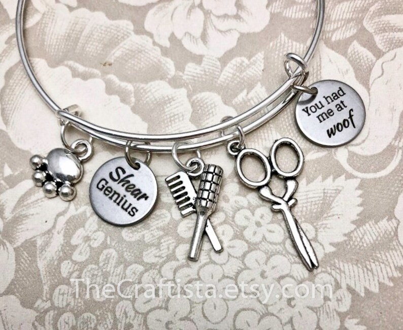 May include: Silver charm bracelet with five charms: a paw print, a comb and brush, a pair of scissors, a charm that says "Shear Genius", and a charm that says "You had me at woof".