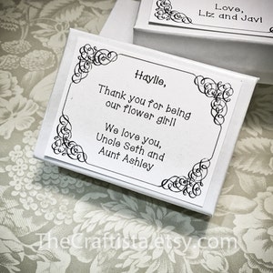 May include: White gift box with a decorative black border and a thank you message for a flower girl. The message reads "Haylie, Thank you for being our flower girl! We love you, Uncle Seth and Aunt Ashley".