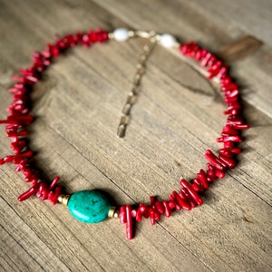 May include: A red coral necklace with a turquoise stone pendant and a gold chain. The necklace is on a wooden surface.