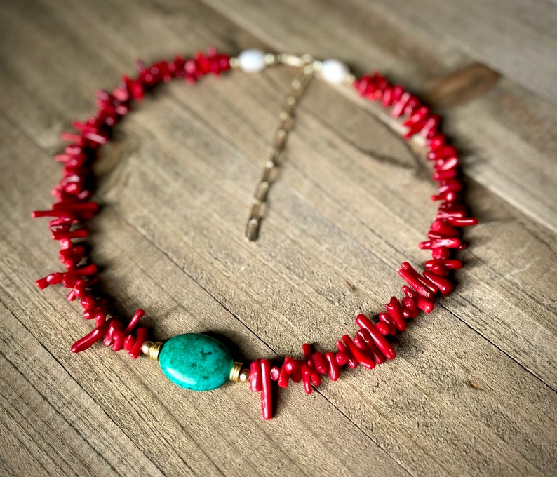 May include: A red coral necklace with a turquoise stone pendant and a gold chain. The necklace is on a wooden surface.